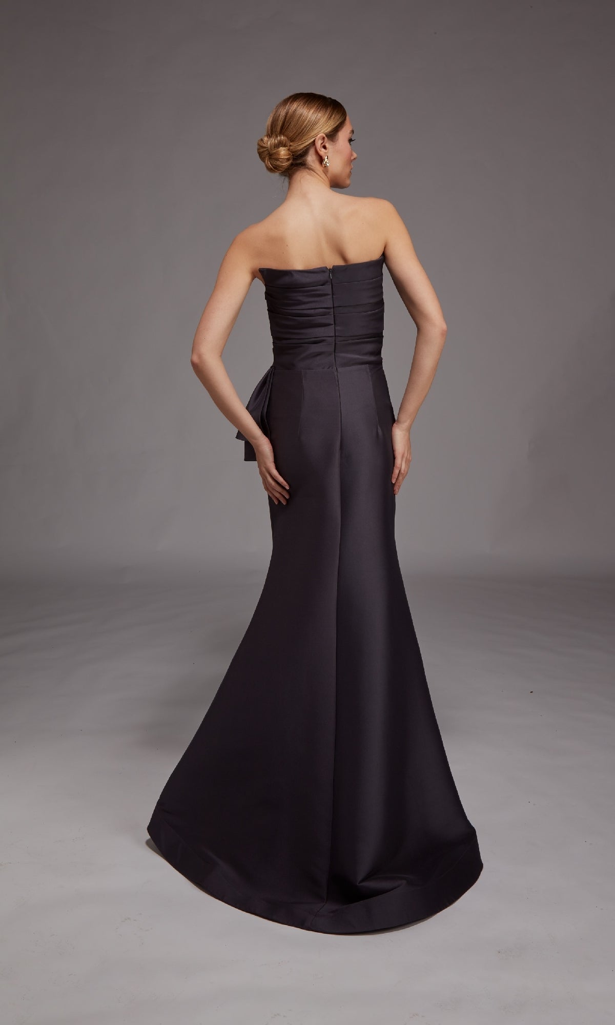 A woman stands facing away, wearing the Alyce 27806 long formal dress—a strapless, floor-length gown with a fitted bodice and flared skirt—her hair styled in a low bun.